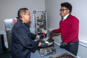 Chef Tello handles a pan over a gas heater as he works with a student on cooking skills. 